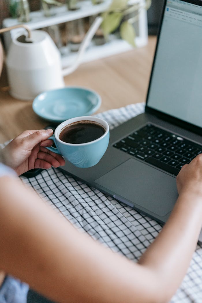 From above of crop unrecognizable female using netbook and drinking mug of coffee at home